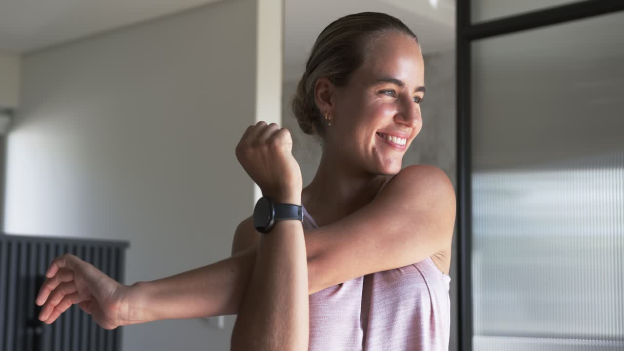 Mujer estirando el brazo izquierdo sobre el pecho con la mano derecha, calentando en casa mientras lleva un reloj inteligente.