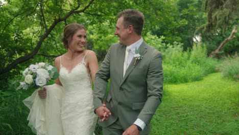 bride and groom walk hand-in-hand through forest clearing, smiling, laughing, and playfully interacting