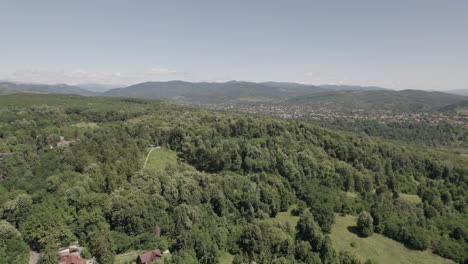 ascending aerial view over dense forest landscape with houses far in the distance