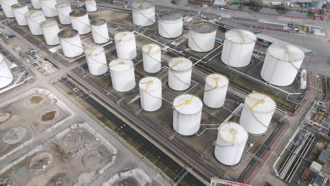 Top-down aerial of tightly clustered white fuel silos with walkways and pipework along a riverside depot, illustrating petrochemical storage logistics