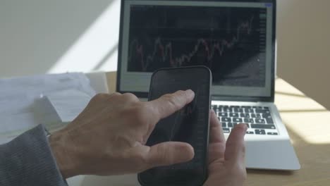 Elder-caucasian-hands-check-stock-market-graphics-in-phone-laptop-vintage-sunny-light-in-living-room-desktop