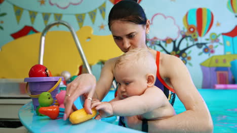 smiling brunette woman holding a plump blond baby in the swimming pool. lovely kid chooses the toy to play with in water.