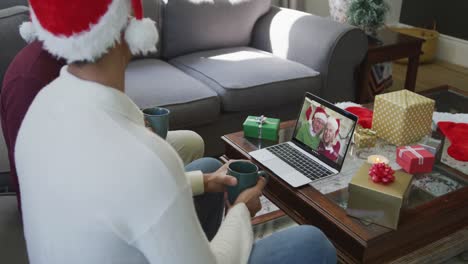 Biracial-father-and-son-with-santa-hats-using-laptop-for-christmas-video-call-with-couple-on-screen