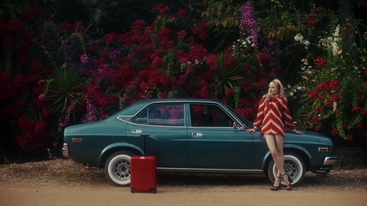 Donna alla moda che posa con auto d'epoca e valigia rossa accanto a fiori vivaci