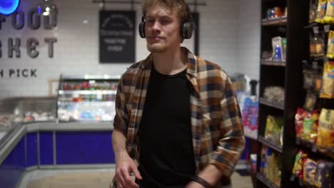 Cheerful-caucasian-guy-with-headphones-walks-through-goods-section-of-the-store-and-listening-to-the-music,-and-funny-dance-wearing-plaid-shirt.-Front-view.-Slow-Motion