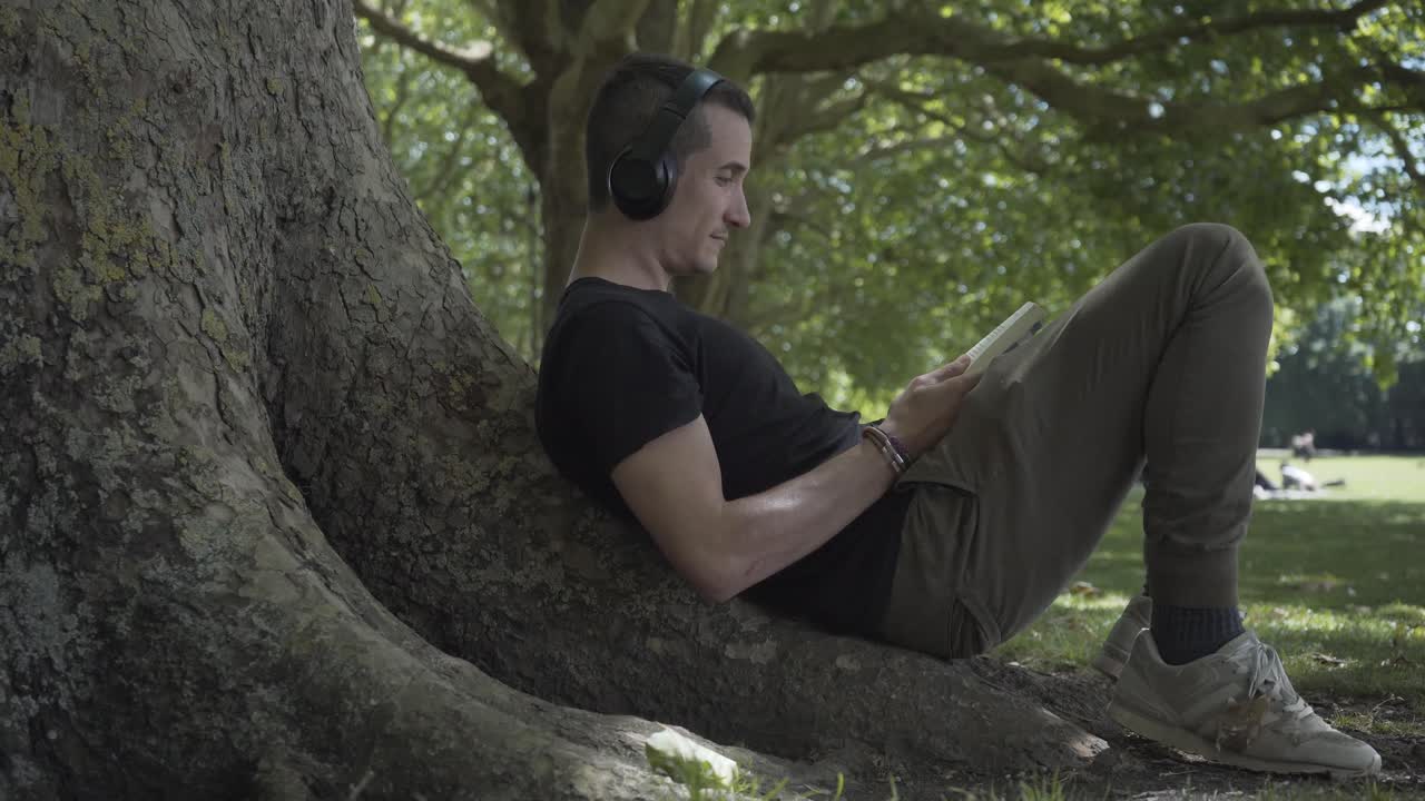joven blanco escuchando música y leyendo un libro está sentado debajo de un árbol disfrutando el momento en un parque de cambridge, inglaterra