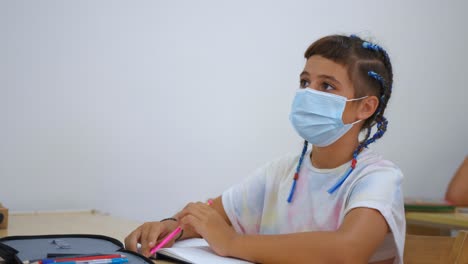 school girl in classroom wearing face mask asking the teacher a question