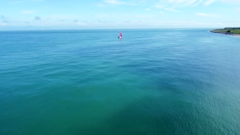 Aerial-chase-of-a-sailboat-along-a-coastline-in-overcast-day-with-varying-sunlight