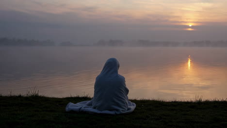 person wrapped in blanket sitting by a lake at sunrise