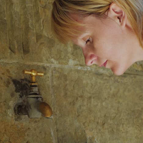 a tired woman washes takes water from an old faucet in a stone wall