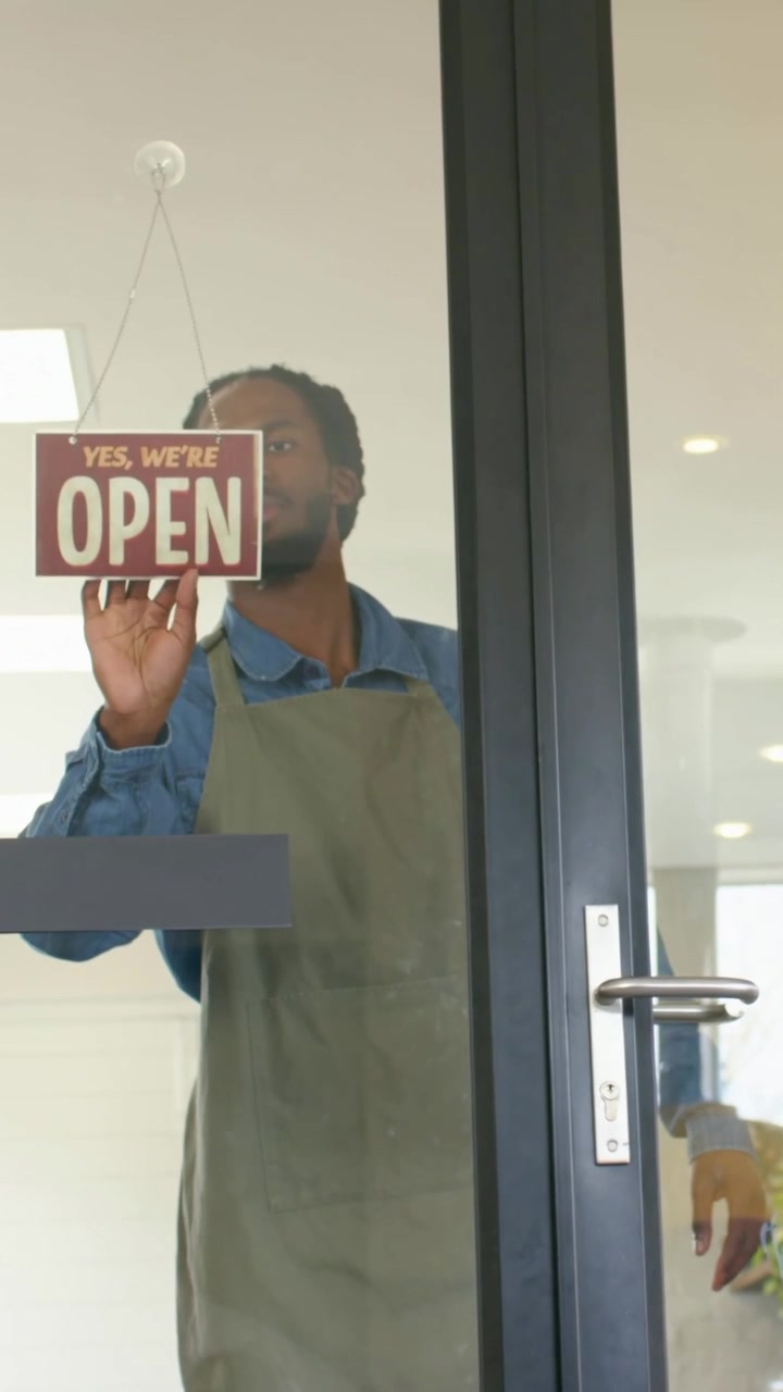 Vertical video: Opening shop aproned worker flipping CLOSED sign to OPEN, unlocking door, exiting