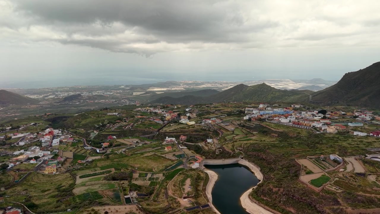 Veduta aerea di un villaggio rurale, un bacino idrico e colline vulcaniche sotto un cielo nuvoloso a Tenerife.