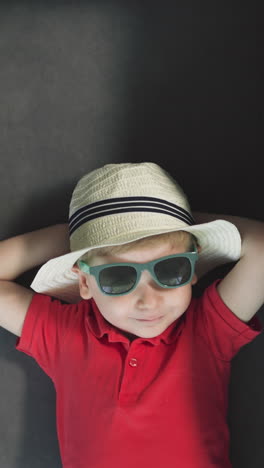 cute toddler with hands behind head rests on couch near black suitcase. small boy in sunglasses and straw hat lies in hotel room upper close view