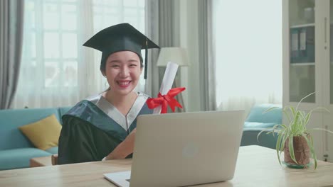 excited asian woman smiling and showing off a university certificate to camera. pretty female graduate wearing a graduation gown and cap sitting on the living room