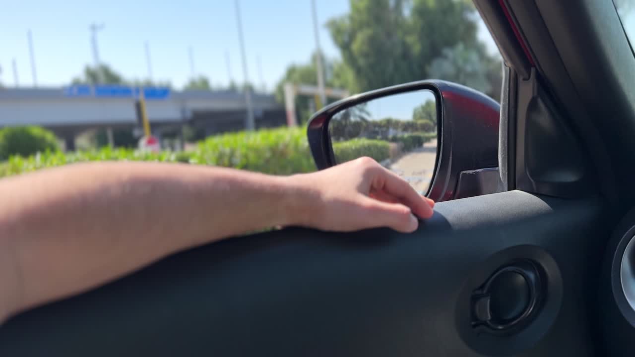 Primo piano di un braccio nudo appoggiato sul pannello della portiera di un'auto con la mano vicino allo specchietto laterale, mentre si guida su un'autostrada urbana soleggiata con alberi verdi e cielo azzurro.