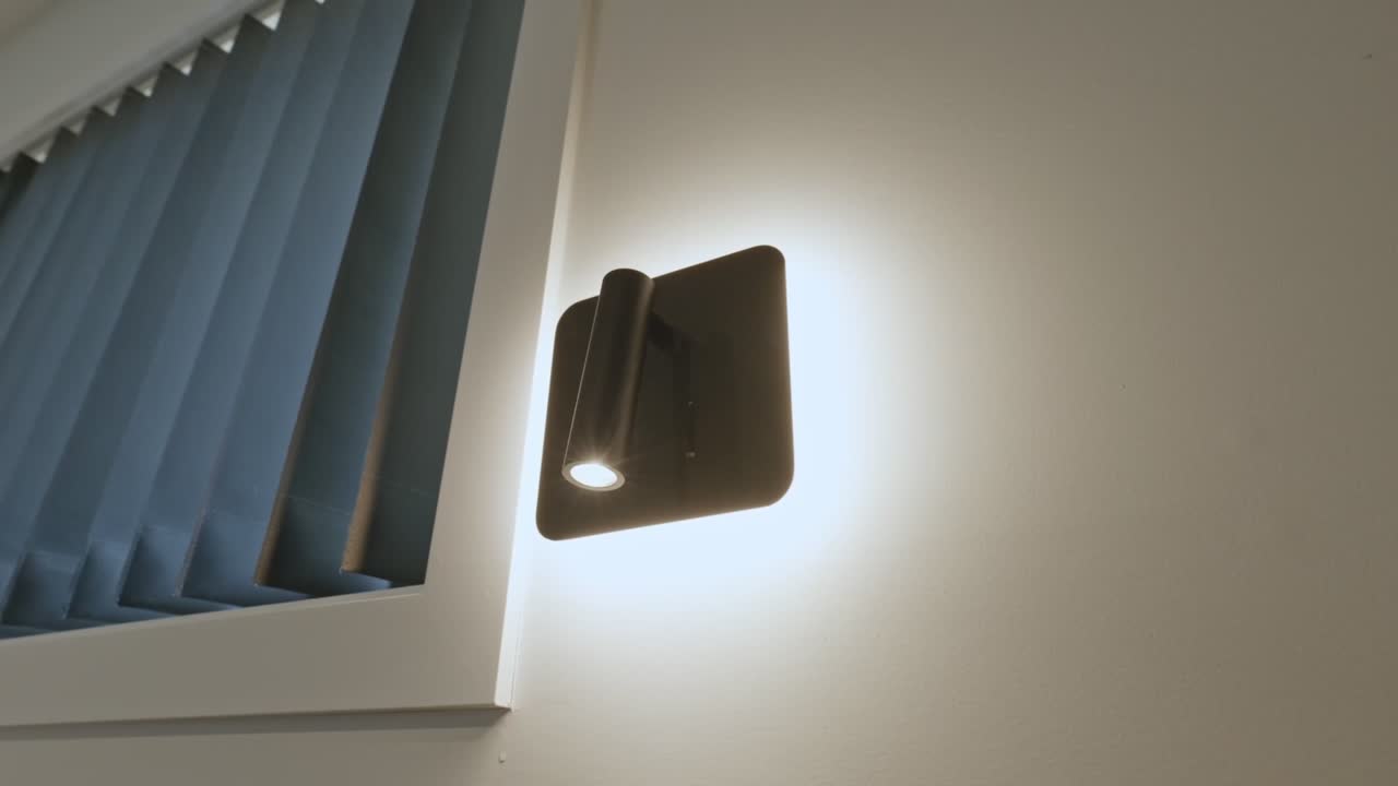 Lavvinklet cirkulær kamerabevægelse omkring en moderne vægmonteret lampe, der kaster et varmt skær på en hvid væg i et beboelsesrum med synlige blå gardiner.