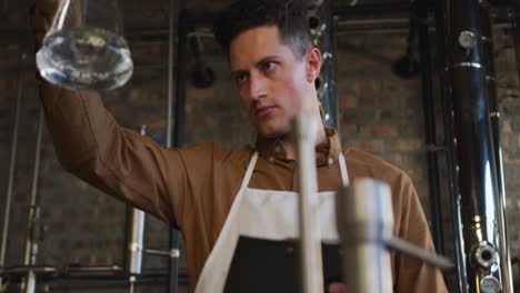 Caucasian-man-working-at-distillery-checking-gin-product-in-flask-and-writing-on-clipboard