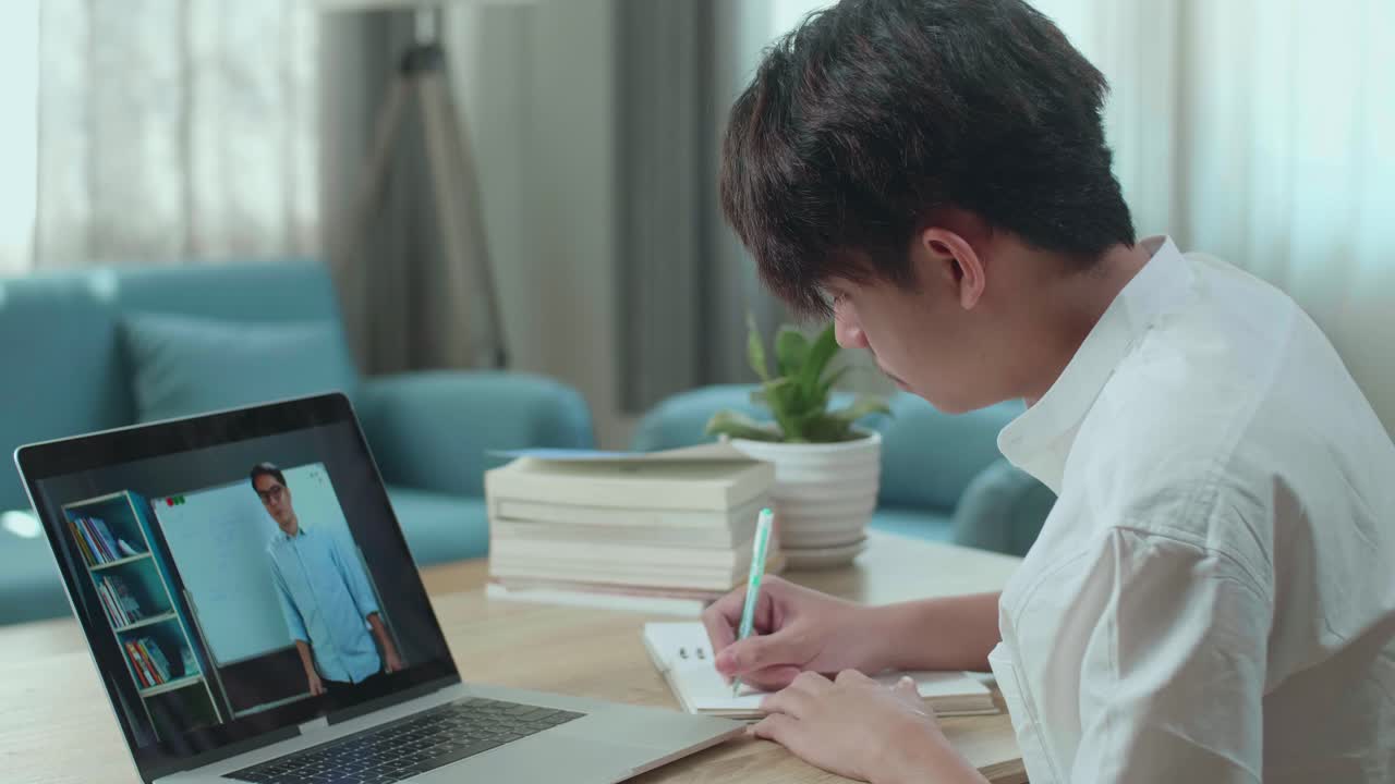 Close Up Of Asian Man Student Look At Teacher In Computer Screen And Write In The Notebook While Studying Online At Home