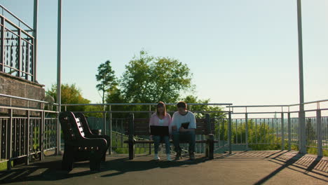 high school students studying outdoors, sitting on benches, one using a laptop and the other holding a tablet, surrounded by railings, trees, and sunlight, creating a calm learning atmosphere