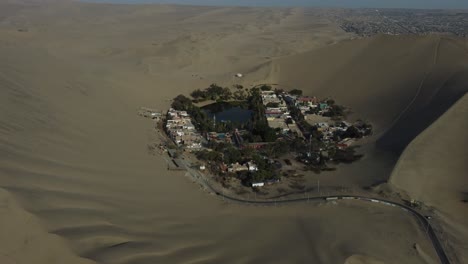 aerial footage of an oasis in the desert. a lagoon surrounded by sand dunes. drone orbits to the right and tilts camera up. located in a village called "huacachina" in ica, peru.