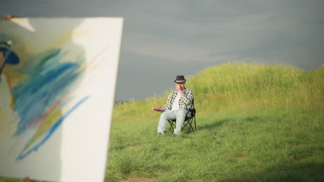 in a sunlit meadow, a focused female artist in a white dress paints on a canvas with a palette in hand, while a man in a black hat and checkered shirt sits calmly in a chair, observing the scene