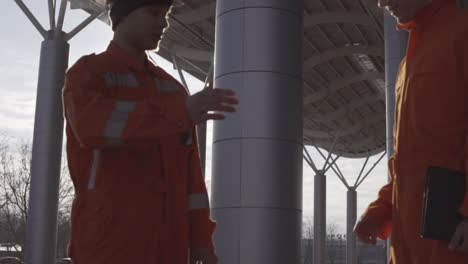 Two-construction-workers-in-orange-uniform-and-hardhats-meeting-each-other-at-the-building-object-and-shaking-hands