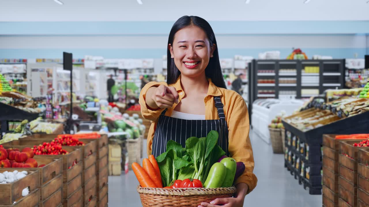 Agricultora asiática sonriendo con una cesta de verduras y haciendo un gesto honesto en una tienda de comestibles