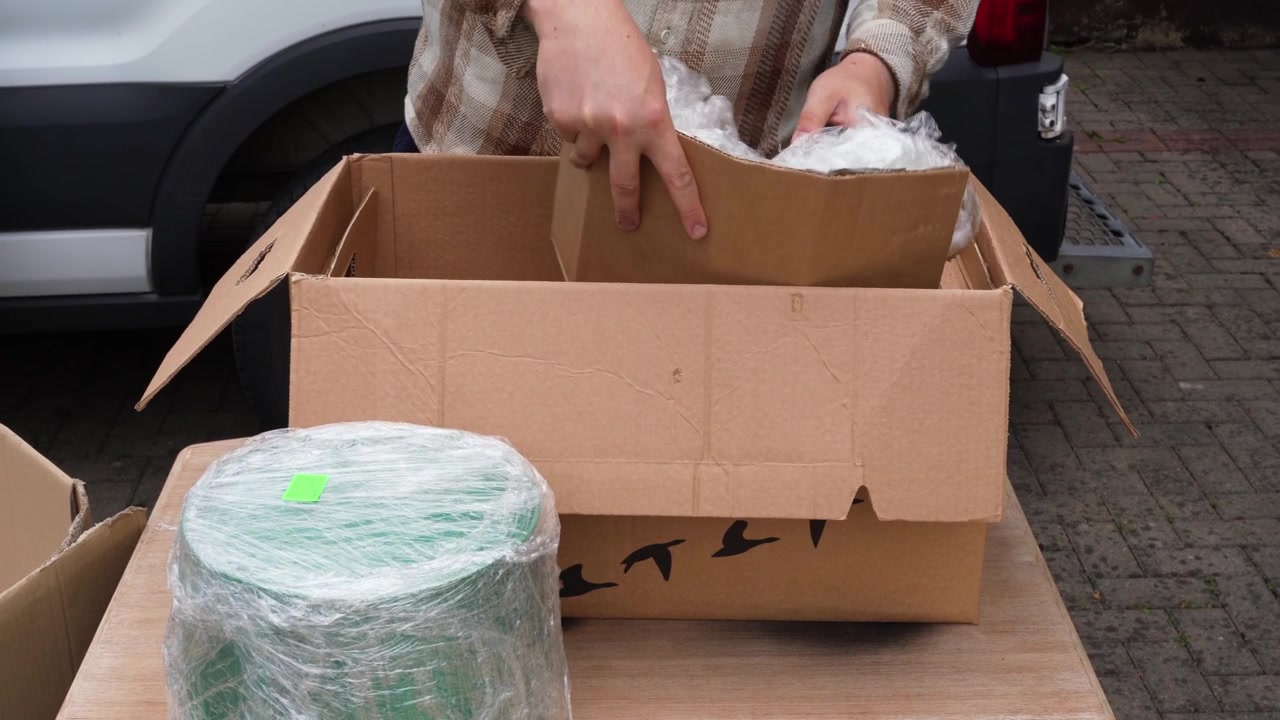 Mani sistemano materiali di imballaggio riutilizzati all'interno di una scatola per pacchi su un tavolo all'aperto, mostrando l'evasione degli ordini e-commerce ecologica
