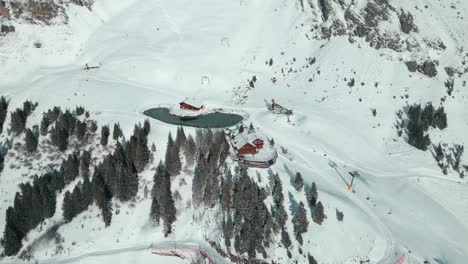 Blick-Von-Oben-Auf-Eine-Herannahende-Drohnenaufnahme-Einer-Hütte-Mit-Einem-Künstlichen-See,-Einer-Kabelhaltestelle-Am-Schneebedeckten-Alpenhang-In-Engelberg,-Brunni,-In-Bahnen,-Schweiz