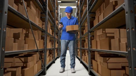 full body of asian male courier in blue uniform using smartphone while delivering a carton in warehouse