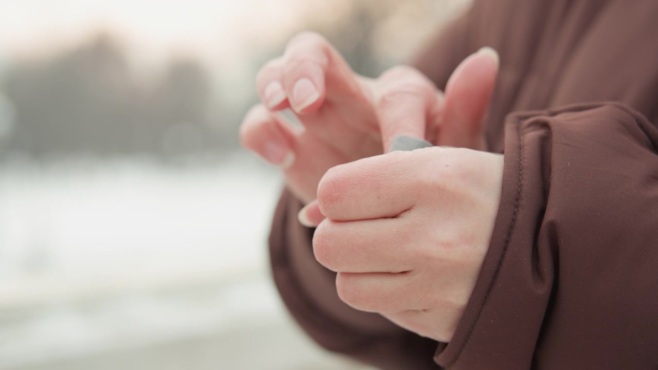 Hands in brown coat pinch, rub, and smooth soft gray cloth near chest height in blurred cold outdoor setting, showing careful tactile movement, gentle adjustment, and natural everyday handling