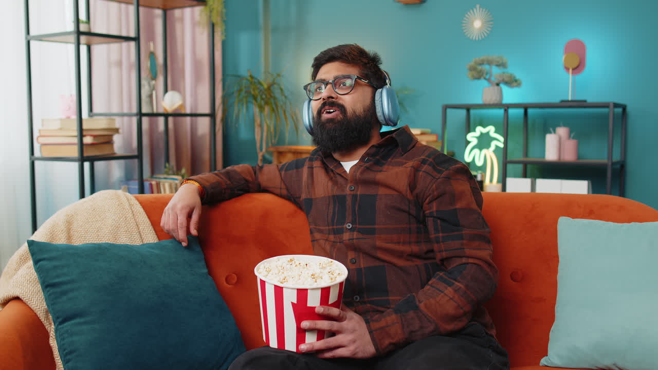 Relaxed arabian man eating popcorn watching movie enjoying film during weekend sitting on home sofa