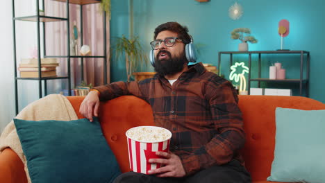 relaxed arabian man eating popcorn watching movie enjoying film during weekend sitting on home sofa