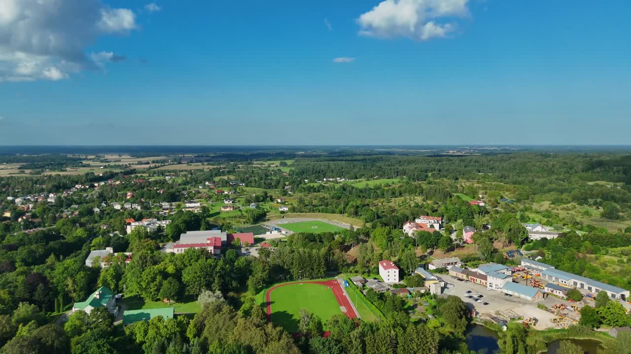 Aerial view of Talsi, Latvia with Green landscape revealing municipal stadium, residential buildings, verdant trees surrounding calm water body under bright sunlight, camera tilting down