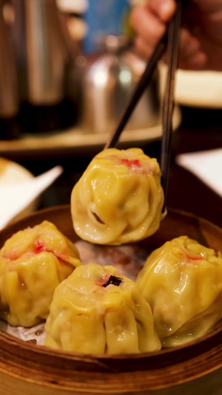A hand uses black chopsticks to lift a steaming dumpling from a bamboo basket.