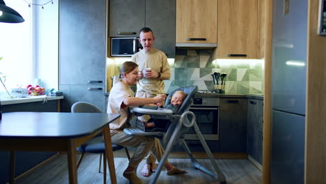 young caucasian family in the kitchen. caring mom feeding her baby and father is watching his beloved ones.
