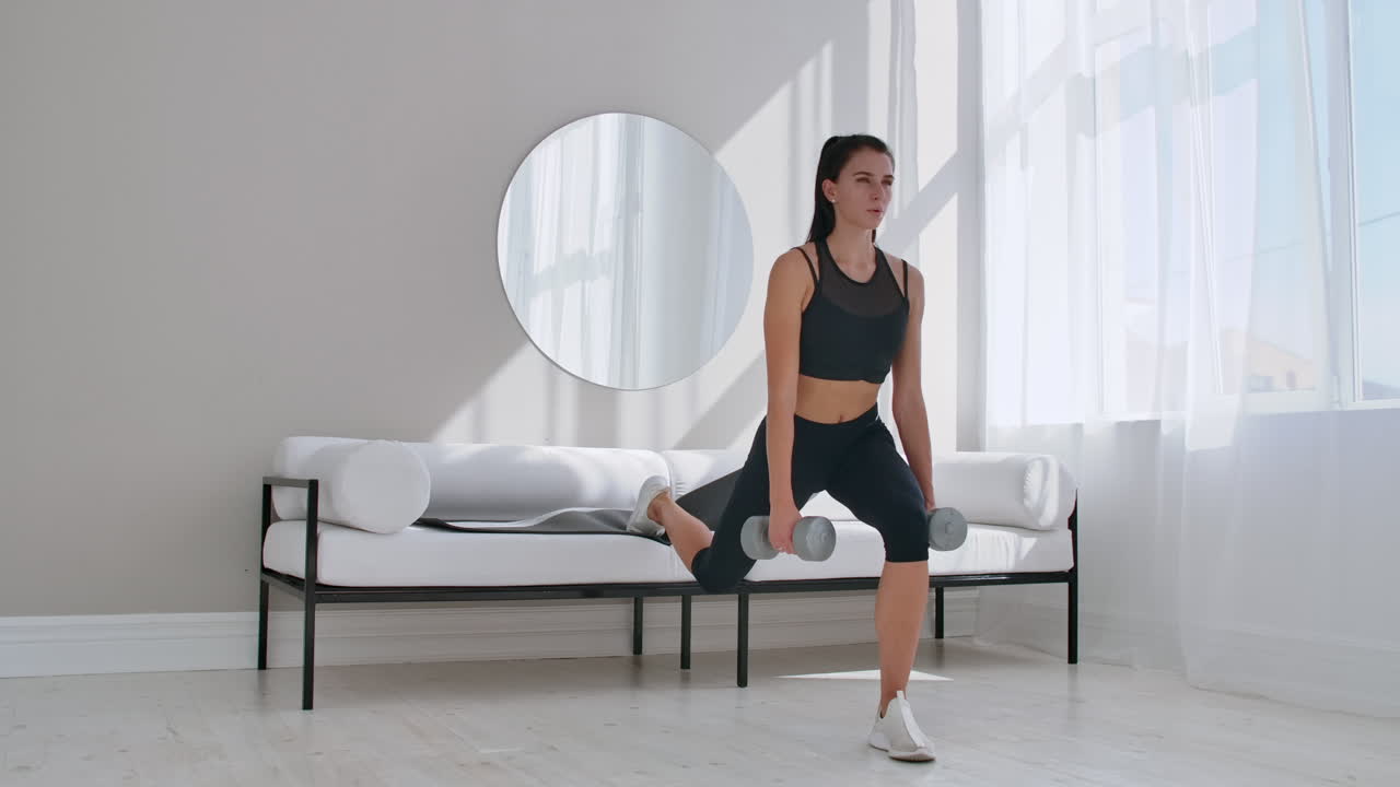 Una bruna in abbigliamento sportivo nero in un appartamento bianco esegue uno split squat con manubri tra le mani, appoggiando il piede sul divano.