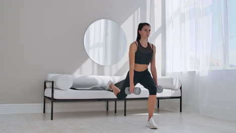 brunette in black sportswear in a white apartment makes a split squat with dumbbells in her hands leaning on the sofa with her foot.