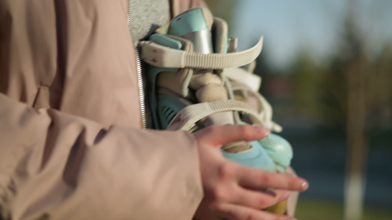 Close-up of a girl's hand carrying a skateboard while walking through a park on a sunny day. The focus is on the hands and skateboard, with the face not visible