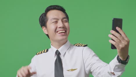 녹색 화면 배경 스튜디오에 서 있는 동안 스마트폰에서 비디오 통화를 하는 아시아 남자 조종사의 손을 흔들고
