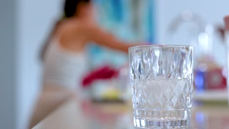 a woman washes dishes in a well-lit kitchen, with focus on a glass in the foreground