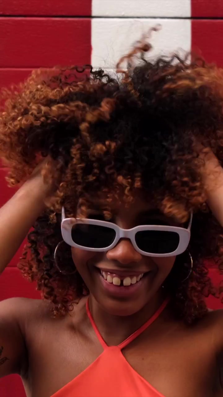 Smiling Young Woman with Curly Hair and Sunglasses Against a Red and White Wall