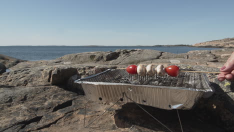 skewers with cherry tomatoes and mushrooms being put onto a disposable bbq grill on the rocks by the ocean side on a sunny day