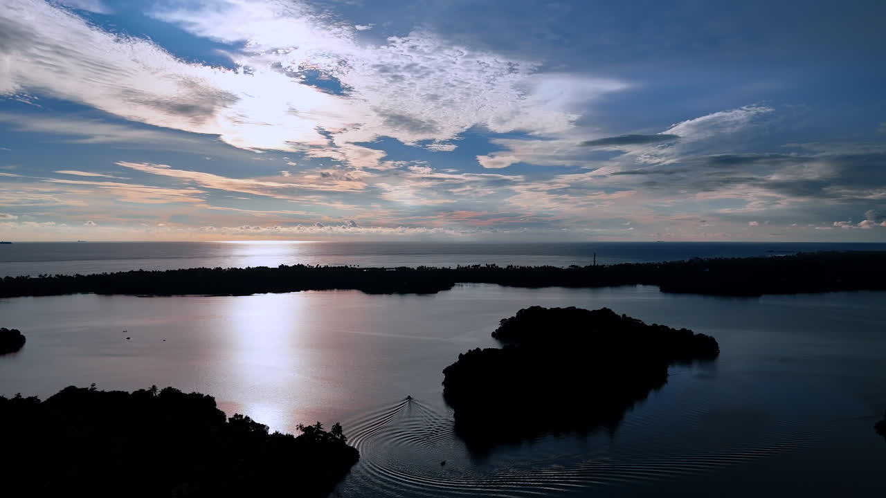 Boat floats by the calm surface of the river at twilight time. Dark silhouettes of bushes surround the waterscape. Light clouds cover the sky at dusk.