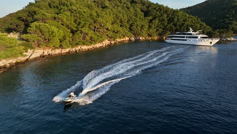 jet ski speeds past anchored white yacht in calm bay with forested slopes and rocky shorelines, kornati islands, croatia