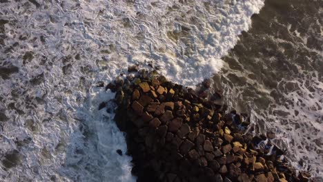 Toma-Aérea-De-Arriba-Hacia-Abajo-De-Olas-Tormentosas-Del-Océano-Que-Se-Estrellan-Contra-Un-Embarcadero-Rocoso-Durante-La-Puesta-De-Sol---Toma-Circular