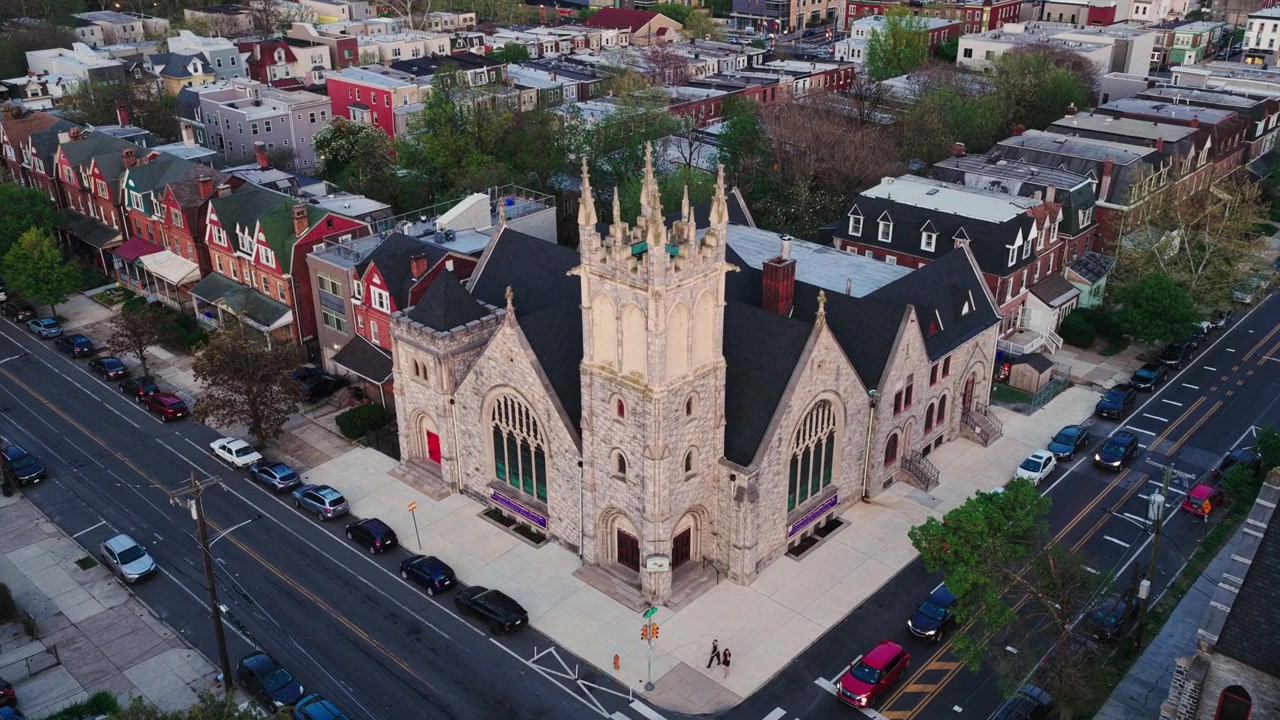 Philadelphia'da bir köşe arsada bulunan, renkli sıra evler, Viktorya dönemi evleri ve ağaçlarla çevrili mahalle sokaklarıyla çevrili görkemli bir Gotik taş kilisenin havadan drone görüntüsü.