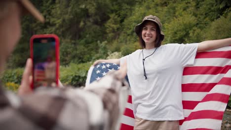 A-blonde-girl-adjusts-and-says-How-to-stand-up-a-brunette-girl-in-a-white-t-shirt-with-an-American-flag-for-a-better-photo-against-the-backdrop-of-the-forest