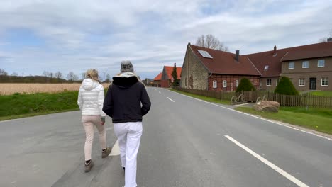 slow motion tracking shot of two adult girls walking along road next to rural wheat field and farm during sunny day with clouds