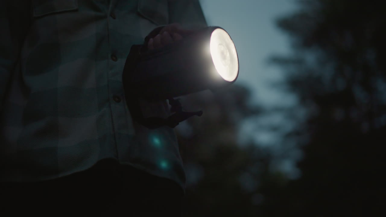 Fascio di luce di una torcia che illumina una foresta oscura, escursionista bianco che perlustra il sentiero con una grande torcia, camicia a quadri e spallaccio dello zaino visibili, cielo crepuscolare tra i pini, atmosfera tesa e silenziosa e movimento concentrato